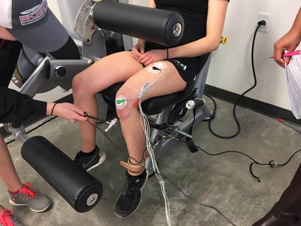 photo of test subject seated and about to be struck with a reflex hammer. The subject's ankle is strapped to a device with a load cell that will measure the force of the reflexive movement.