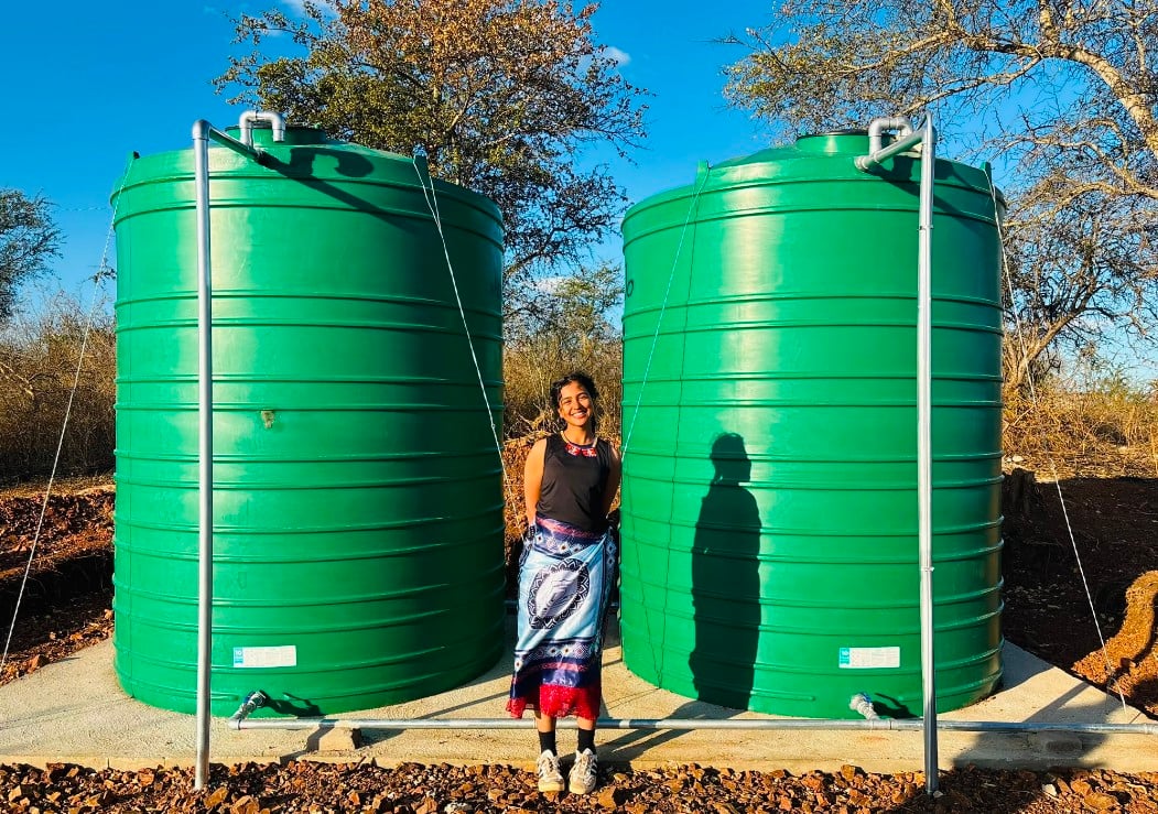 A woman in a black tank top and printed skirt between two green water tanks about twice her height in an outdoor setting