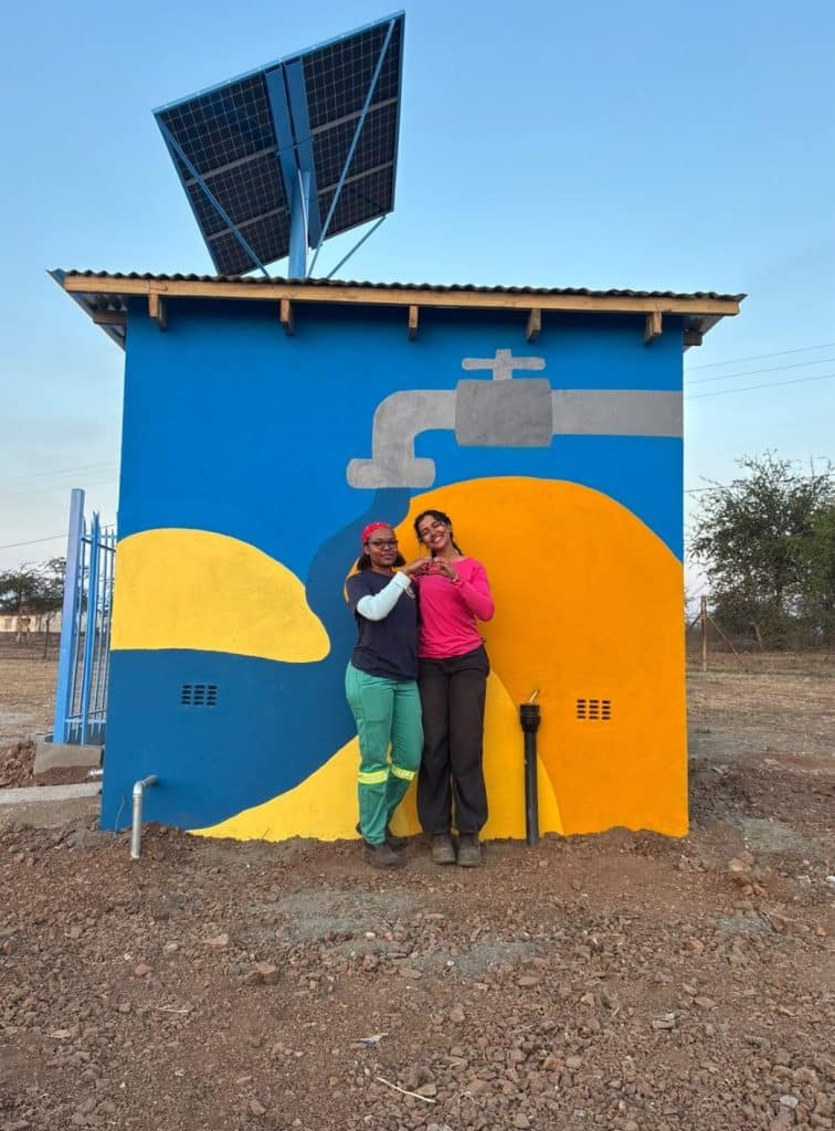 two women in frint of a small building with a painted image on the wall behind them. The image is of a water spout emitting colors