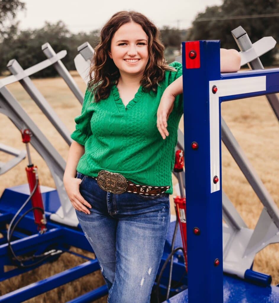 Closeup of a woman in a green shirt and jeans standing on a blue and gray hay bale trailer in the background.