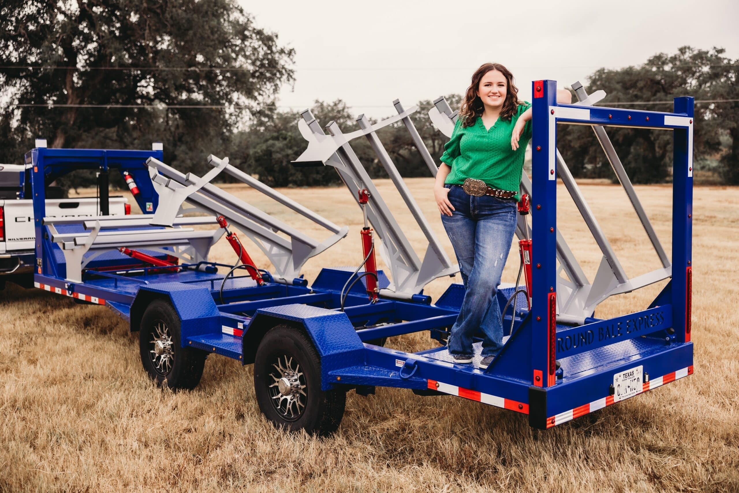Picture of a woman in a green shirt and jeans standing on a blue hay bale trailer with gray hinged bale cradles at various degrees of pivot. They pivot along the far side of the trailer. The trailer looks like a flat bed with four wheels, two on each side and spaced in the middle third of the flat bed. Only the wheels in the foreground are visible.