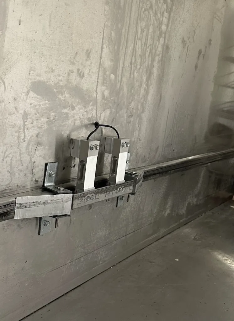 photo of closeup of installed load cells in food waste measurement system. it shows ramps that guide load bearing wheels onto a thin metal bar attaching two load cells at their load point. This bar completes the wheel track where the load cells are positioned.