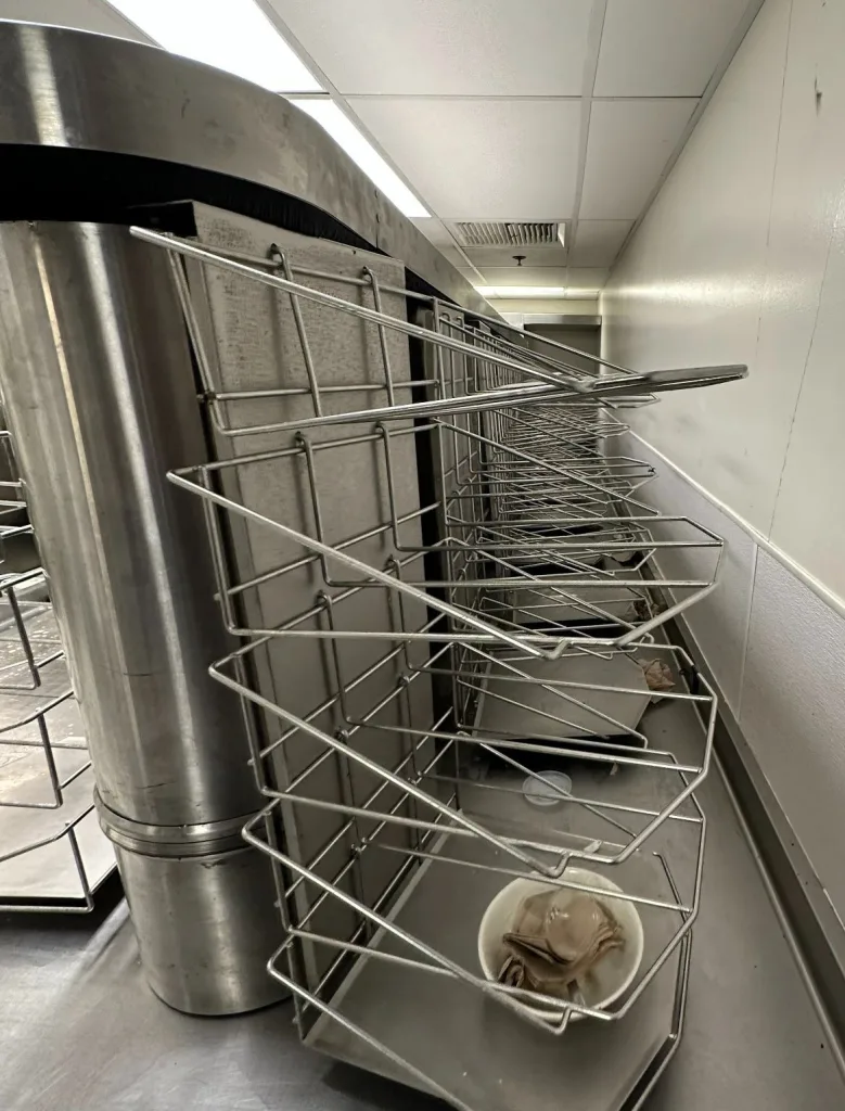 Photo of kitchen view of the University of the Pacific cafeteria tray return rotating system. It shows racks that support returned trays stacked horizontally, one tray wide. These racks are supported along the back by a vertical, inverted L sheet metal component that attaches to a track along the top of a support wall via pins at the top endpoint of the inverted L. Wheels aligned horizontally along the back of this sheet metal roll along a track 2/3 of the way down from the top of the inverted L. These guide the racks and prevent the sheet metal from scraping along the wall when the racks are loaded.